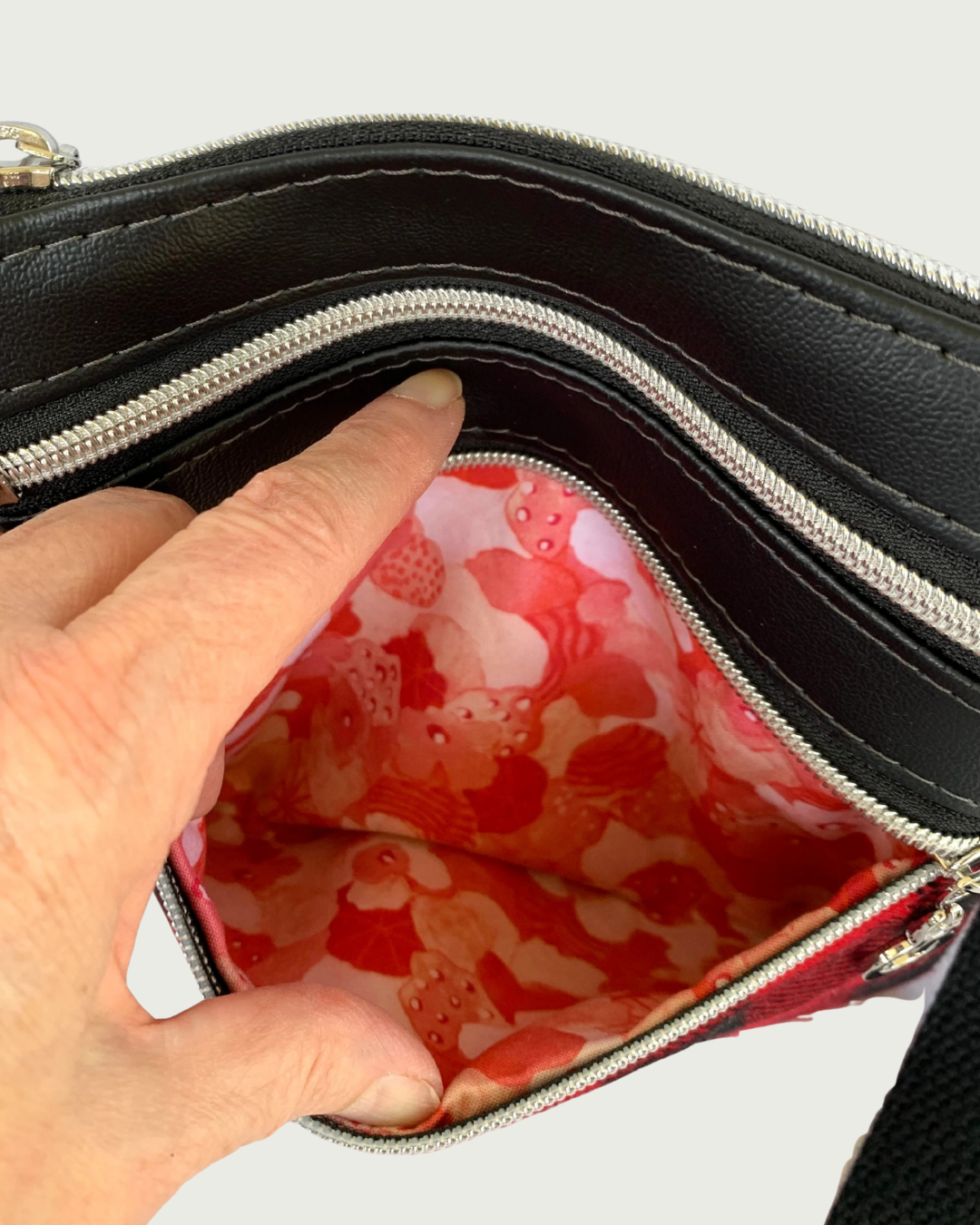 An open nickel zippered pocket of a handmade crossbody bag of black faux leather held open by a hand revealing a red, pink, and white floral cotton lining. Above that is another closed zipped pocket and a top zippered closure,