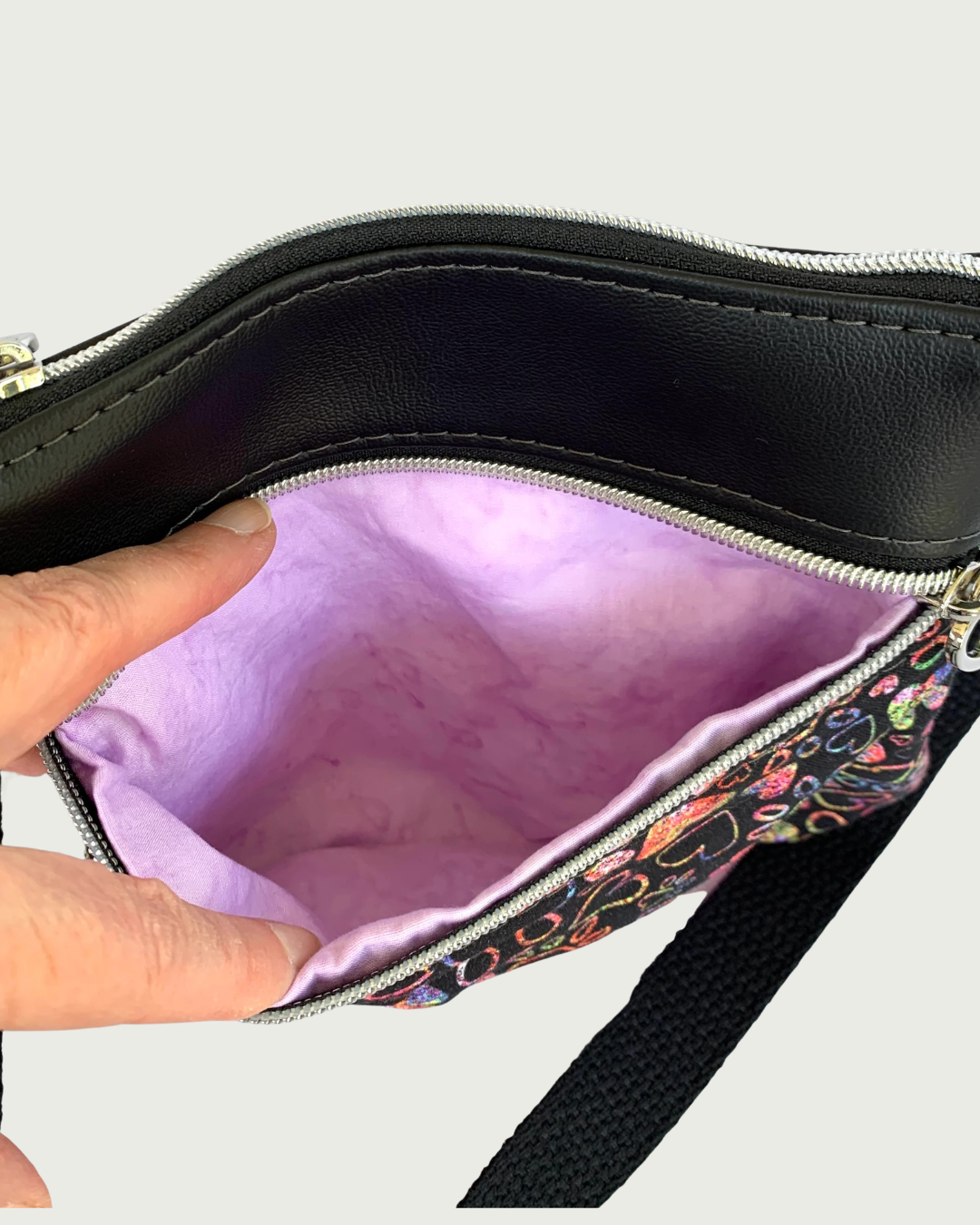 Handcrafted crossbody bag of black fabric with colorful hearts and paw prints and black faux leather accents. An adjustable and detachable shoulder strap of black cotton webbing. Fingers are holding open a zippered pocket, revealing the lavender cotton lining. Above that is a top zippered closure,