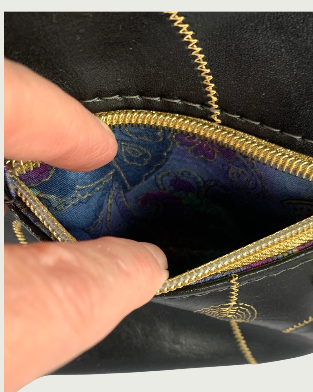 Handcrafted black vinyl slouchy shoulder bag with closeup view of two fingers holding open a small gold zipper showing the interior lining of a pocket in multicolor floral pattern with gold thread running through.