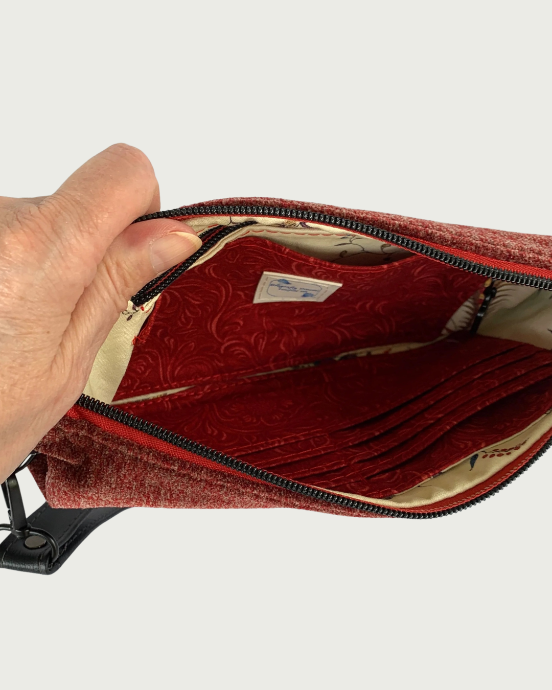 A hand holding open a handmade merlot chenille-like polyester wristlet wallet revealing an open a slip pocket in front of a zippered pocket with a Dragonfly Dreams label sewn in on one side and eight buit-in card slots with a slip pocket behind it on the other side.