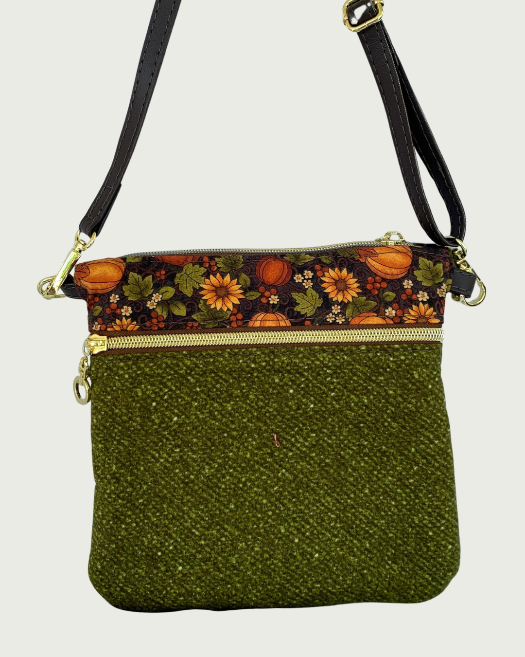 Handmade olive green crossbody bag with a textured fabric body, a zippered pocket featuring a pumpkin and sunflower patterned band between above it, gold-tone hardware, a top zippered closure, hanging by a thin, coffee adjustable and detachable shoulder strap.