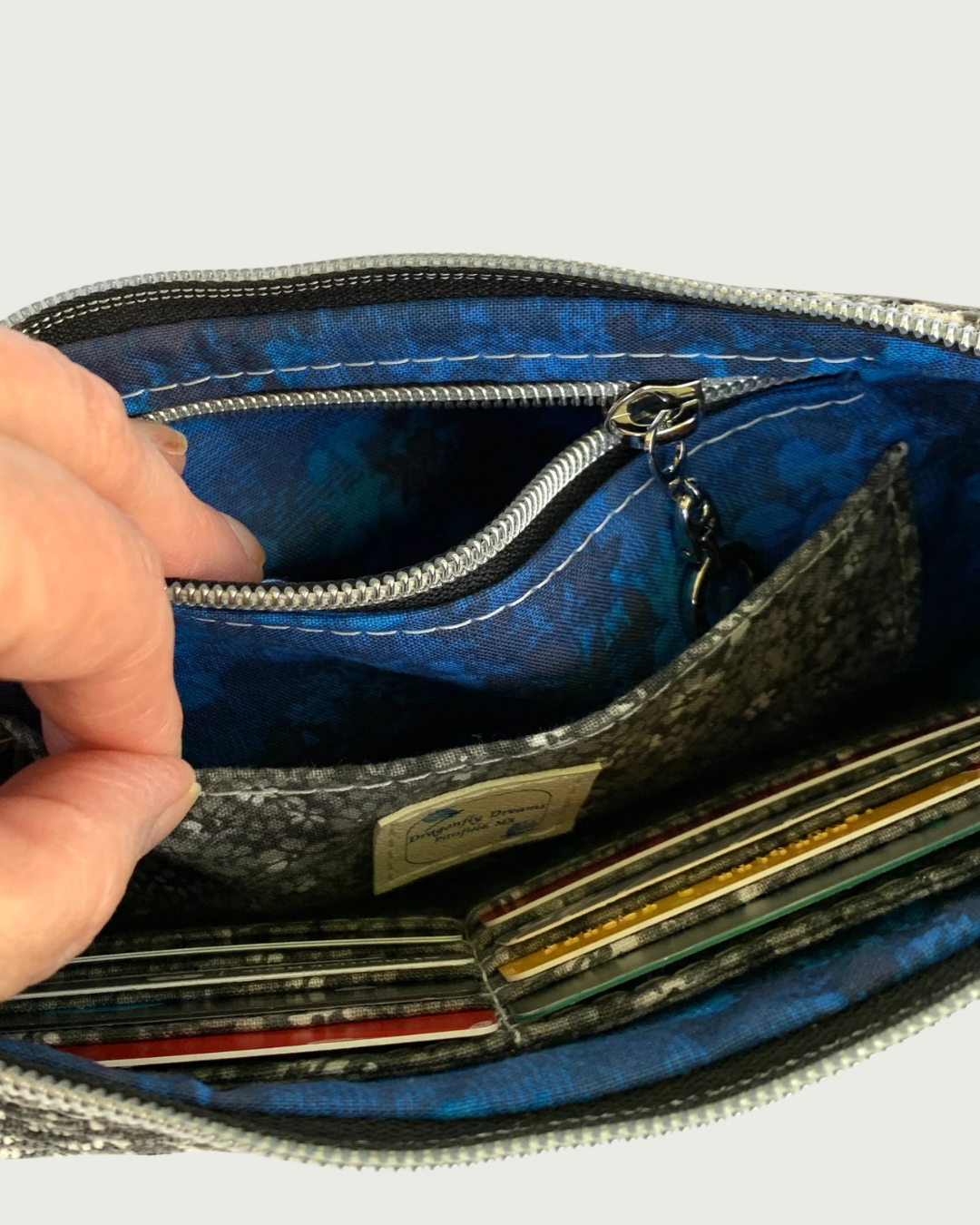Close-up of a zippered wallet or card case opened by a hand, showing a blue patterned interior lining, gunmetal zipper, and card slots with cards inserted.