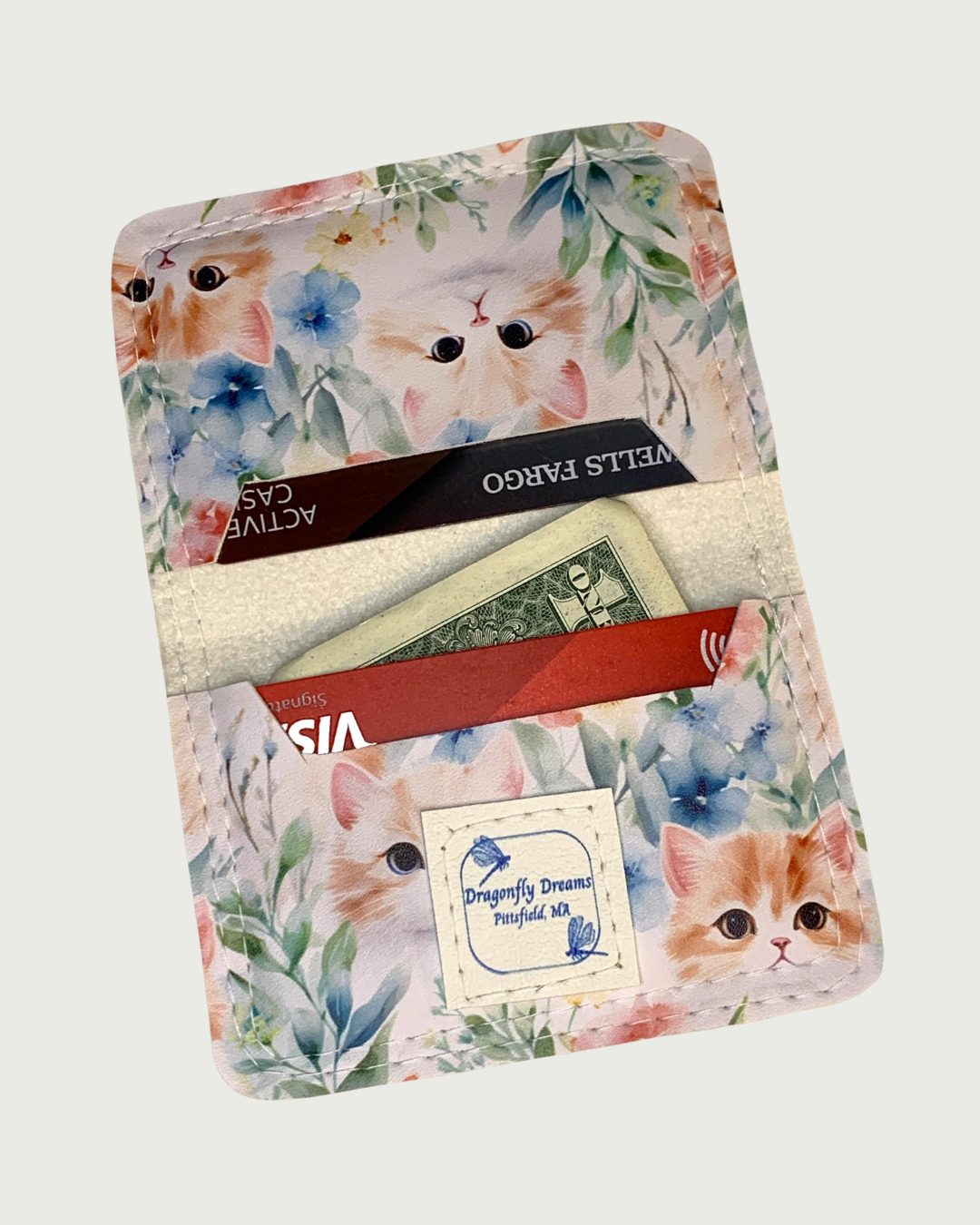Handcrafted compact bifold wallet made from white faux leather vinyl printed with sweet kitten heads popping out of a flower garden. This is the inside view of the wallet open displaying cash and cards in both slots. A Dragonfly Dreams label is sewn onto the bottom card slot.