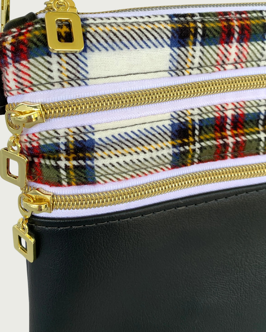 Closeup view of handmade crossbody bag of black faux leather and red, black and white classic plaid cotton flannel. Two zippered pockets have gold-tone open square pulls, gold-tone zipper teeth, and white zipper tape. The open square pull from the top zippered closure is showing.