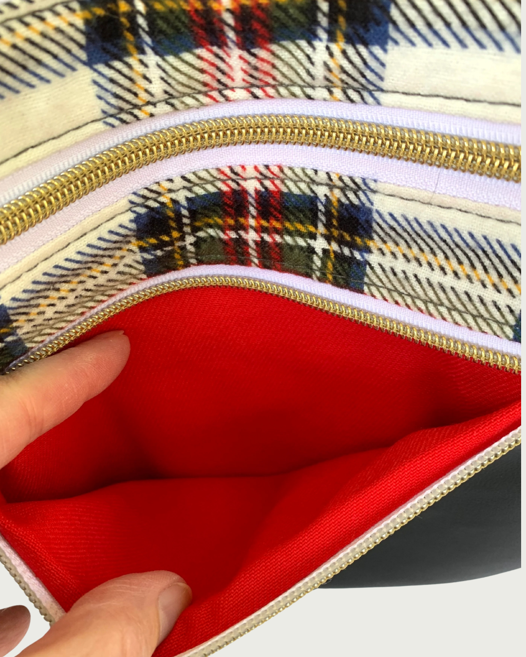 An open zippered pocket of a handmade crossbody bag of classic red, black, and white cotton plaid flannel with black faux leather accents, being held open by a couple of fingers revealing a red cotton broadcloth lining. The zipper details of the pocket are gold-tone on white as is the closed zippered pocket above.