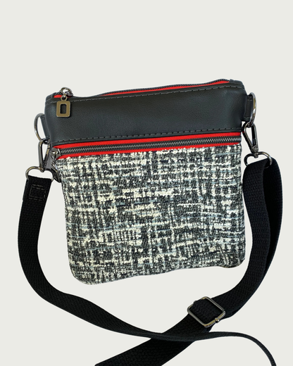 Handcrafted black and white tweed crossbody bag with black faux-leather accents, gunmetal hardware throughout including zipper details on top closure and pocket, and a black adjustable, detachable cotton webbing shoulder strap, shown on a light gray background.