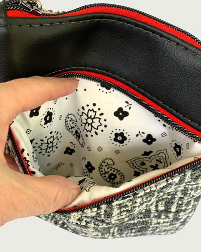 An open zippered pocket of a handmade crossbody bag of black and white tweed with black faux leather accents, held open by a hand revealing a black on white printed cotton lining. The zipper details of the pocket are gunmetal on red as is the top zippered closure above.