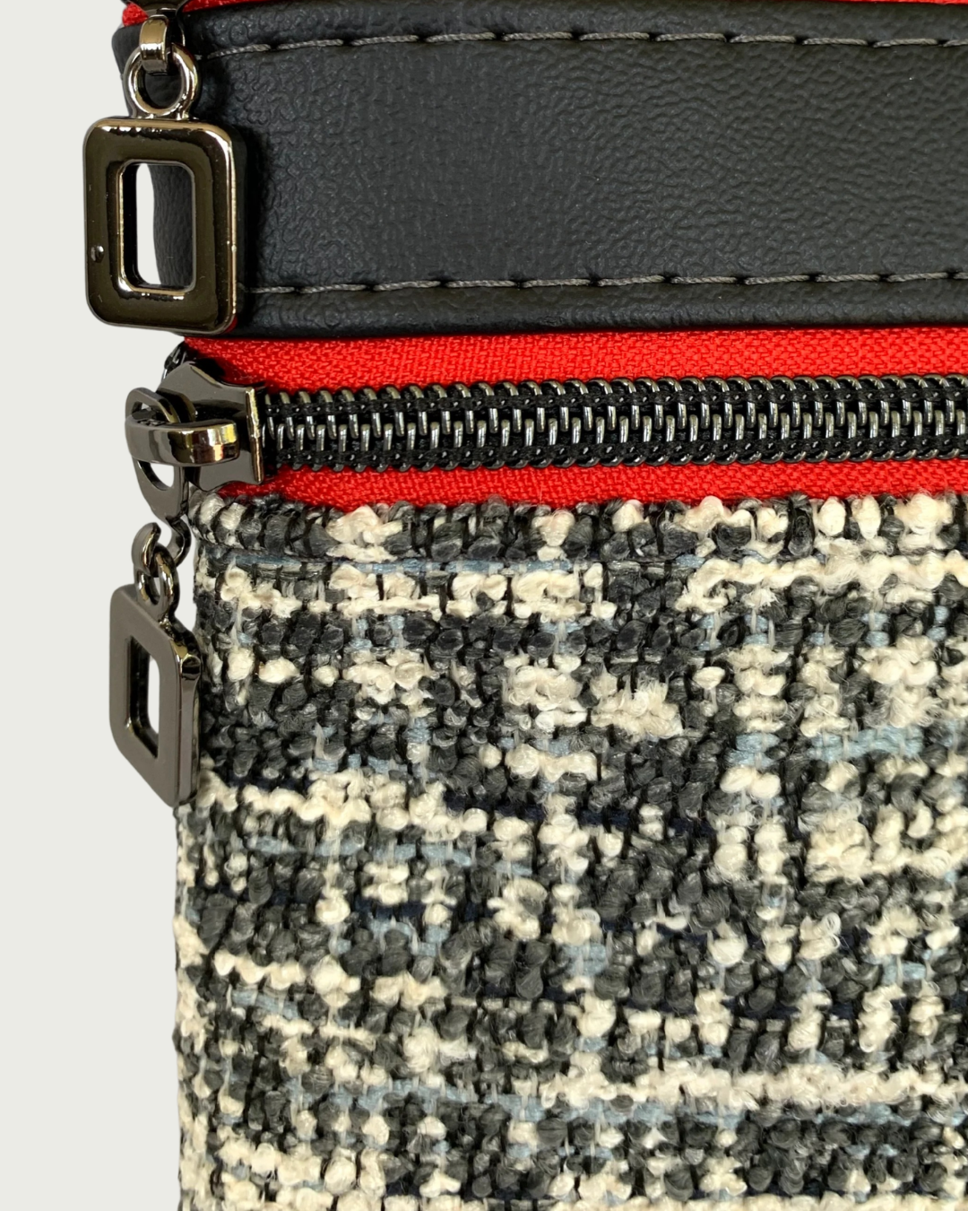 Closeup view of handmade crossbody bag of black and white tweed with black faux leather trim.. Two zippered pockets have gunmetal open square pulls, gunmetal zipper teeth, and red zipper tape.