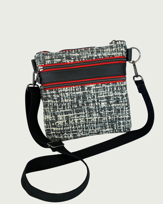 Handcrafted black and white tweed crossbody bag with black faux-leather accents, gunmetal hardware throughout including zipper details on top closure and two pockets, and a black adjustable, detachable cotton webbing shoulder strap, shown on a light gray background.