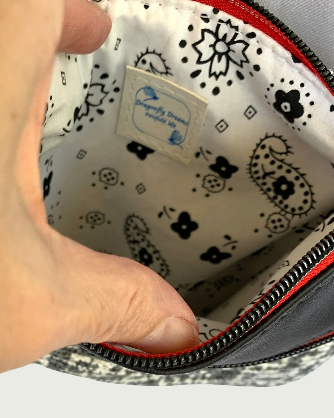 Interior view of Handmade crossbody bag of black and white tweed fabric revealing a black on white printed cotton lining of paisleys and flowers with a sewn-in Dragonfly Dreams label. The top zippered closure has gunmetal zipper teeth and red zipper tape.