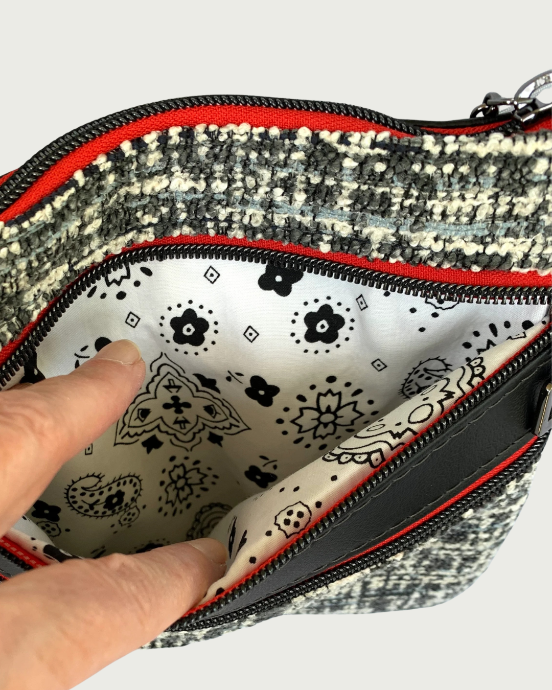 An open zippered pocket of a handmade crossbody bag of black and tweed with black faux leather accents, held open by a hand revealing a black on white printed cotton lining. The zipper details of the pocket are gunmetal on red as is the top zippered closure above and the zippered pocket below.