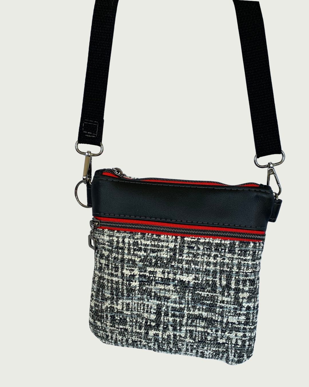Handcrafted crossbody bag of black and white tweed with black faux leather accents, gunmetal zipper details on red zipper tape of top closure and pocket, and a black adjustable. detachable cotton webbing shoulder strap, shown on a light gray background.