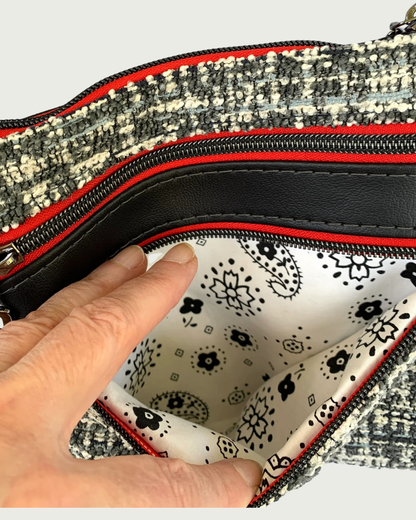 An open zippered pocket of a handmade crossbody bag of black and white tweed with black faux leather accents, being held open by a couple of fingers revealing a black on white printed cotton lining. The zipper details of the pocket are gunmetal on red as is the closed zippered pocket above and the top zippered closure.