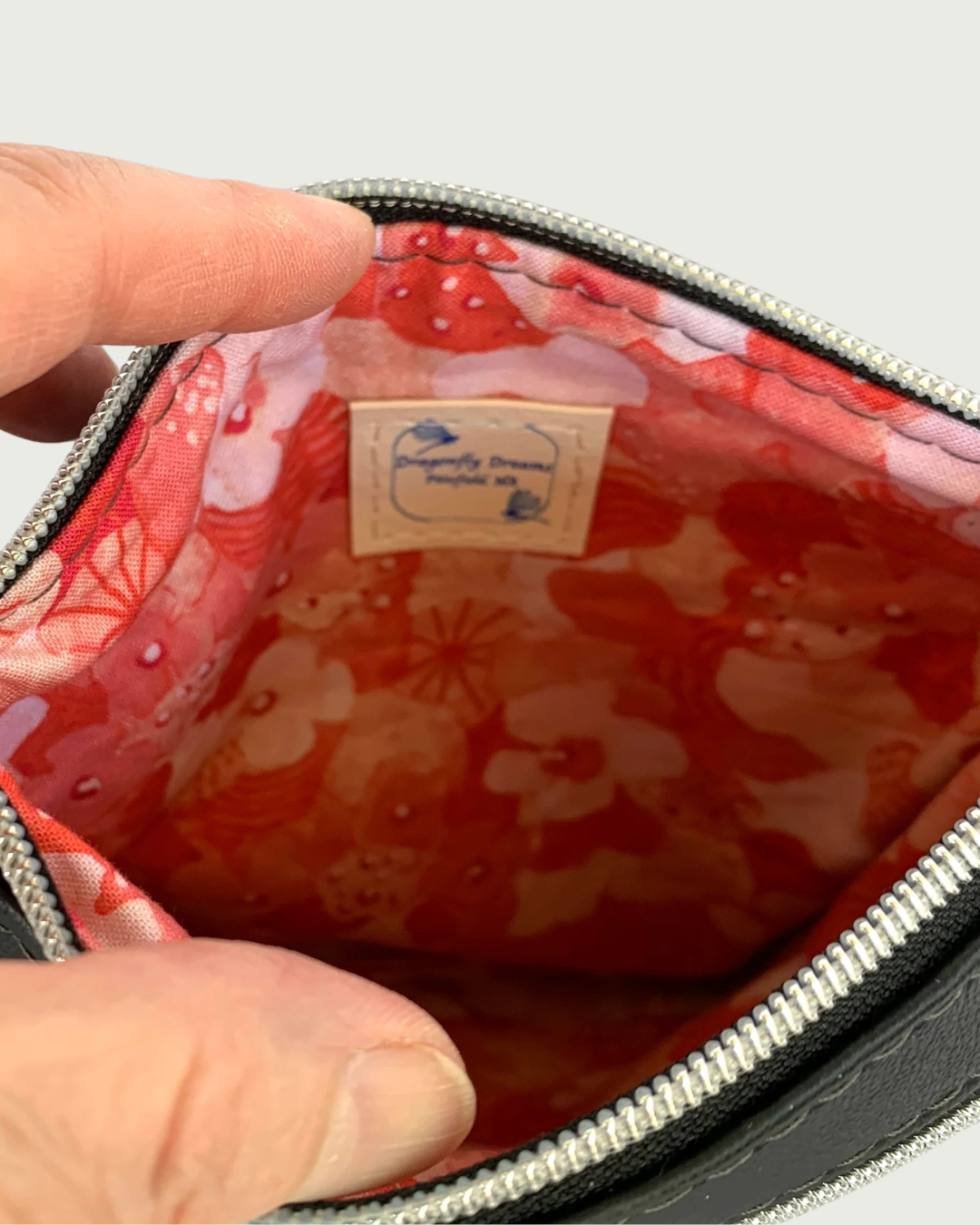 Interior view of Handmade crossbody bag of black faux leather revealing a red, pink, and white floral printed cotton lining with a sewn-in Dragonfly Dreams label. The top zippered closure has nickel-colored zipper teeth and black zipper tape.