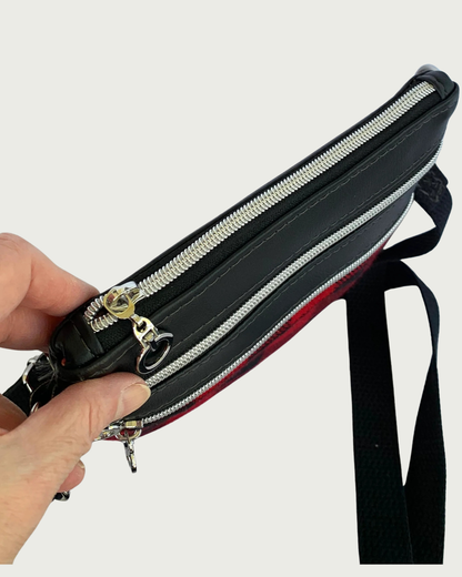 Top view of Handmade crossbody bag of red and black buffalo plaid with black faux leather accents. An adjustable and detachable shoulder strap of black cotton webbing. The top zippered closure and the two zippered pockets have nickel dangling circle pulls, nickel-colored zipper teeth, and black zipper tape.