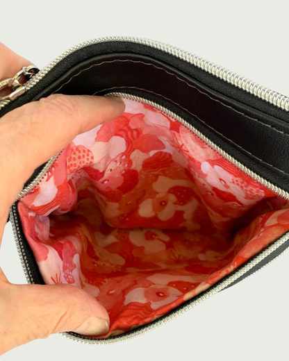 An open zippered pocket of a handmade crossbody bag of black faux leather held open by two fingers revealing a red, pink, and white floral cotton lining. Above that is the top zippered closure, and there is another closed zipped pocket below.