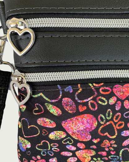 Closeup view of handmade crassbody bag of black fabric with colorful hearts and paw prints and black faux leather accents. Two zippered pockets have nickel heart pulls, nickel-colored zipper teeth, and black zipper tape.