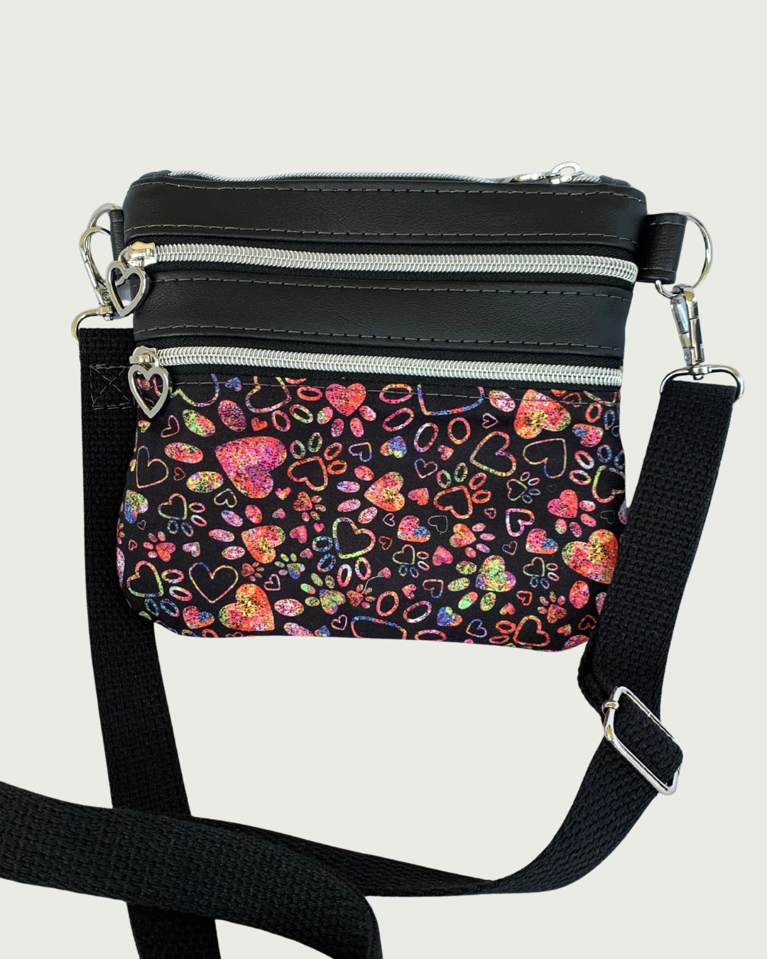 Handmade crossbody bag of black fabric with colorful hearts and paw prints and black faux leather accents. An adjustable and detachable shoulder strap of black cotton webbing. Nickel hardware throughout including heart pulls on the zippered pockets.