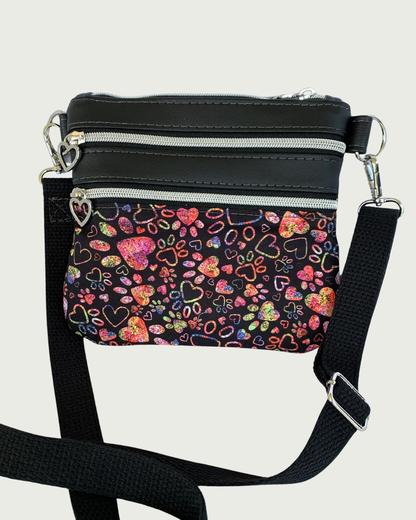 Handmade crossbody bag of black fabric with colorful hearts and paw prints and black faux leather accents. An adjustable and detachable shoulder strap of black cotton webbing. Nickel hardware throughout including heart pulls on the zippered pockets.