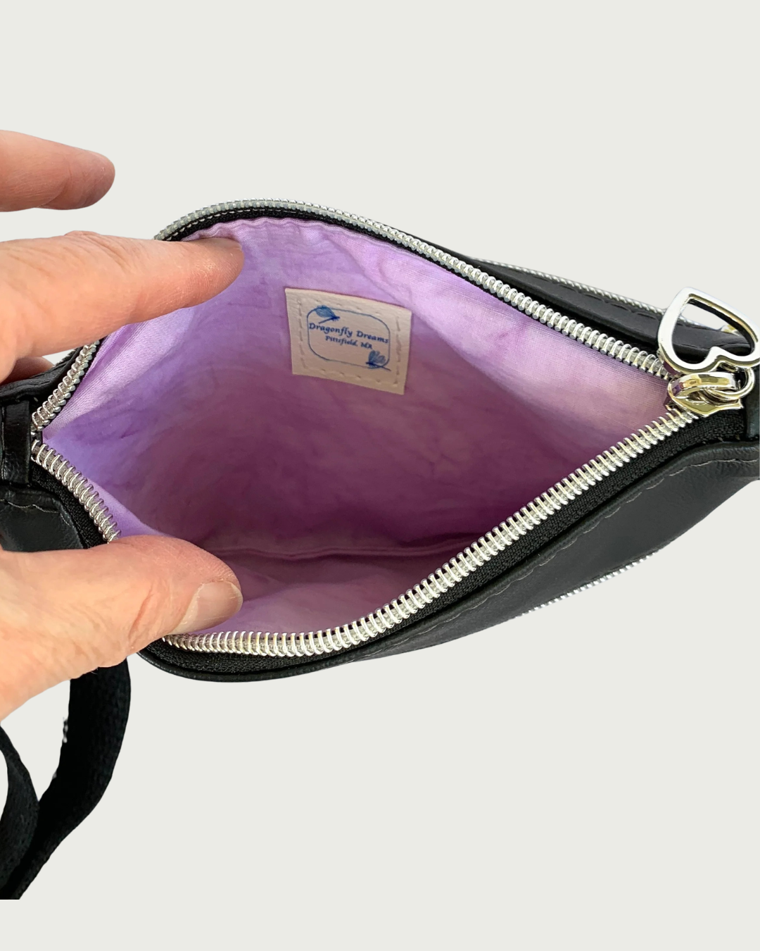 Interior view of Handmade crossbody bag of black faux leather with an adjustable and detachable shoulder strap of black cotton webbing. The lining is a lavender cotton with a sewn-in Dragonfly Dreams label. The top zippered closure has a nickel heart pull, nickel-colored zipper teeth, and black zipper tape.