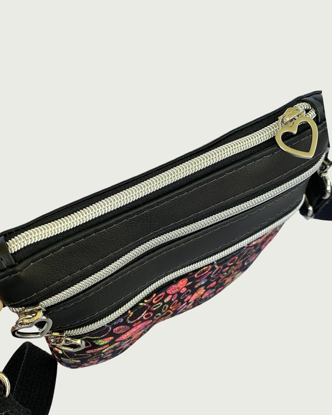 Top view of Handmade crossbody bag of black fabric with colorful hearts and paw prints and black faux leather accents. An adjustable and detachable shoulder strap of black cotton webbing. The top zippered closure and the two zippered pockets have nickel heart pulls, nickel-colored zipper teeth, and black zipper tape.