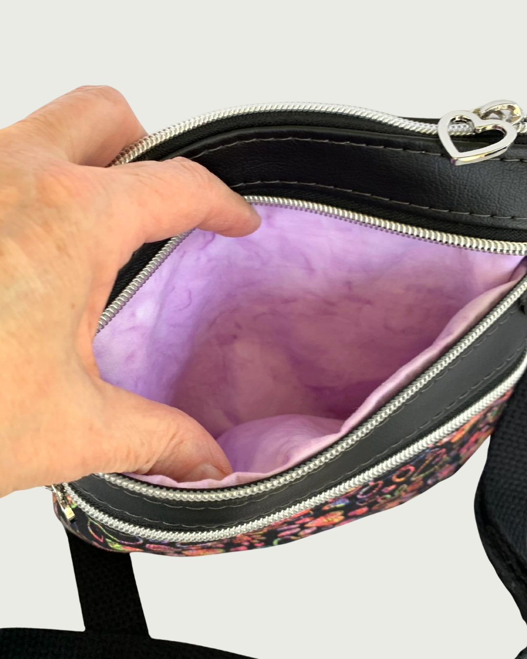 Handmade crossbody bag of black fabric with colorful hearts and paw prints and black faux leather accents. An adjustable and detachable shoulder strap of black cotton webbing. A hand is holding open the top zipper pocket, revealing the lavender cotton lining. Above that is the top zippered closure has a nickel heart pull, and there is another closed zipped pocket below.