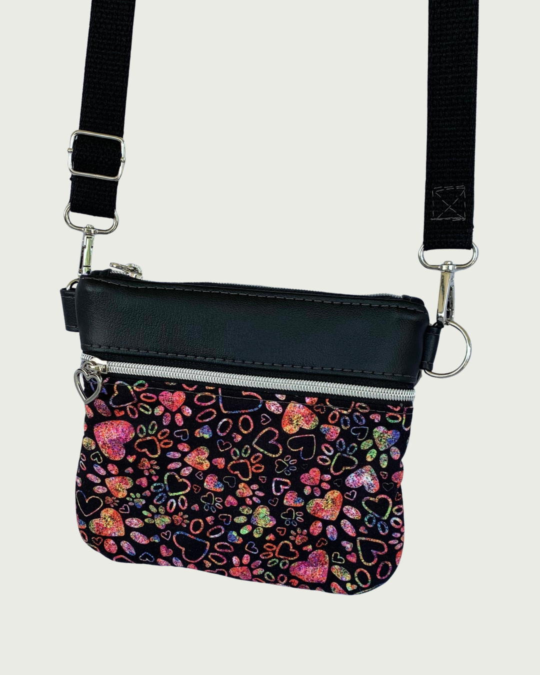Handcrafted crossbody bag of black fabric with colorful hearts and paw prints and black faux leather accents. An adjustable and detachable shoulder strap of black cotton webbing. Nickel hardware throughout including heart pulls on the top closure and the zippered pocket.