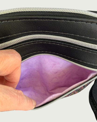 Handcrafted crossbody bag of black fabric with colorful hearts and paw prints and black faux leather accents. An adjustable and detachable shoulder strap of black cotton webbing. A hand is holding open the bottom zipper pocket, revealing the lavender cotton lining. Above that is another closed zipped pocket  and a top zippered closure,