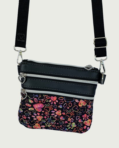 Handcrafted crossbody bag of black fabric with colorful hearts and paw prints and black faux leather accents. An adjustable and detachable shoulder strap of black cotton webbing. Nickel hardware throughout including heart pulls on the top closure and the two zippered pockets.
