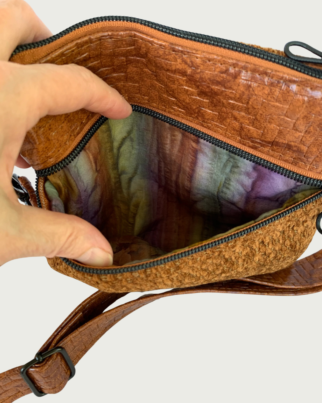 Handmade crossbody bag of brown chenille fabric with brown textured faux leather accents including an adjustable and detachable shoulder strap. Matt black hardware throughout. A hand is holding open the pocket revealing an autumn watercolored striped cotton woven lining.