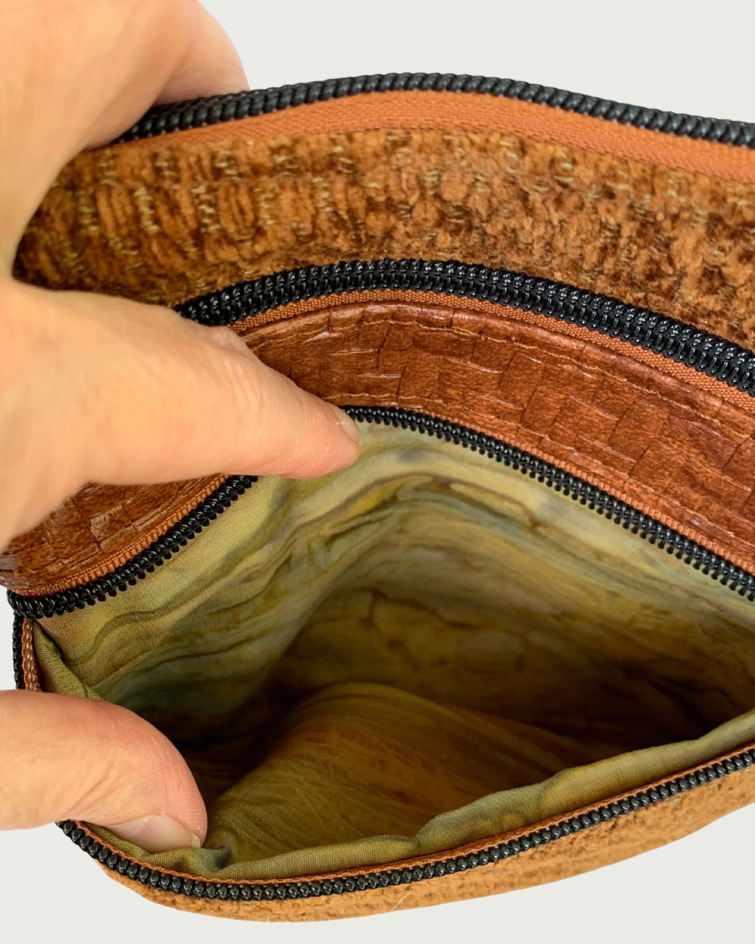 A closeup view of a handcrafted crossbody bag of brown chenille fabric with brown textured faux leather accents. A hand is holding open the pocket revealing an autumn watercolored striped cotton woven lining. Matt black hardware throughout including another zippered pocket and the top zipper closure above it.