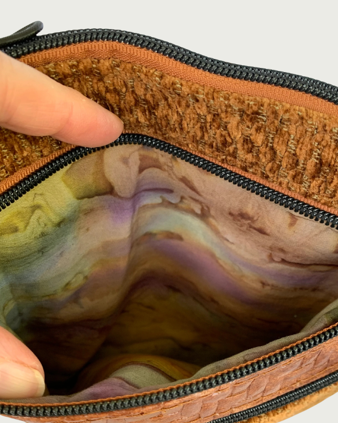 A closeup view of a handcrafted crossbody bag of brown chenille fabric with brown textured faux leather accents. A hand is holding open the pocket revealing an autumn watercolored striped cotton woven lining. Matt black hardware throughout including another zippered pocket below it and the top zipper closure above it.