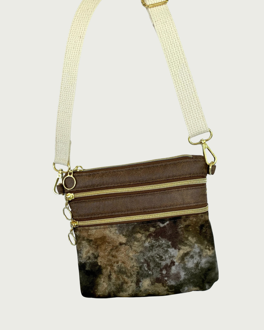 Handmade crossbody bag with camouflage print body and brown, distessed leather accents, two zippered pockets and a top zippered closure with cirlcle zipper pulls, a natural colored cotton webbing adjustable and detachable shoulder strap, and gold-tone hardware throughout.