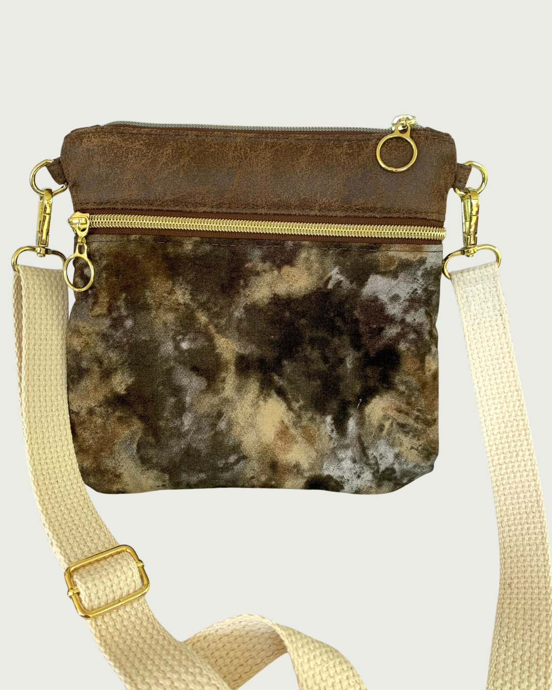 Handcrafted crossbody bag with camo print body and brown, distessed leather accents, a zippered pocket and a top zippered closure with cirlcle zipper pulls, a natural colored cotton webbing adjustable and detachable shoulder strap, and gold-tone hardware throughout.