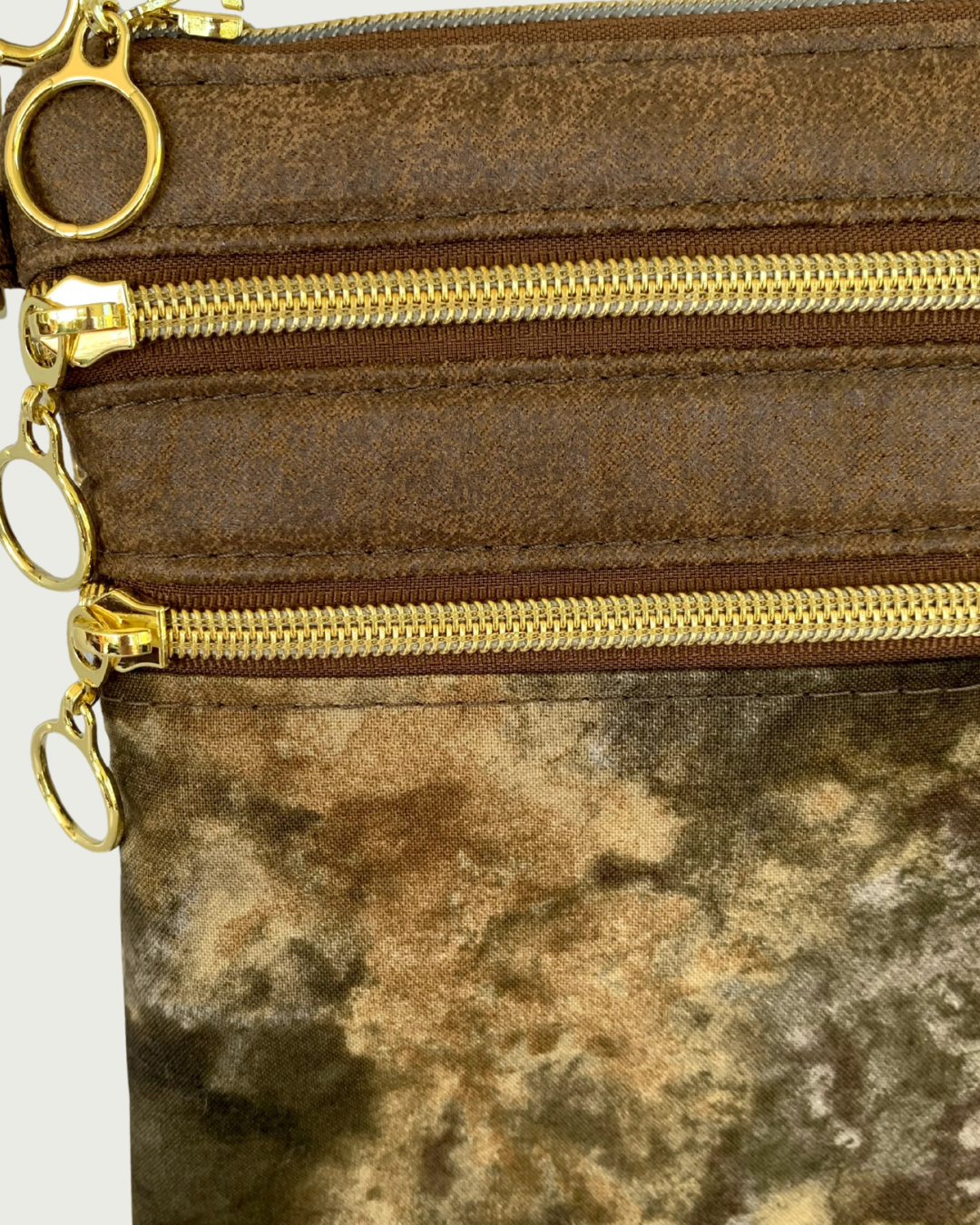 Handcrafted crossbody bag with camo print body and brown, distessed leather accents, two zippered pockets and a top zippered closure with cirlcle zipper pulls closeup view.