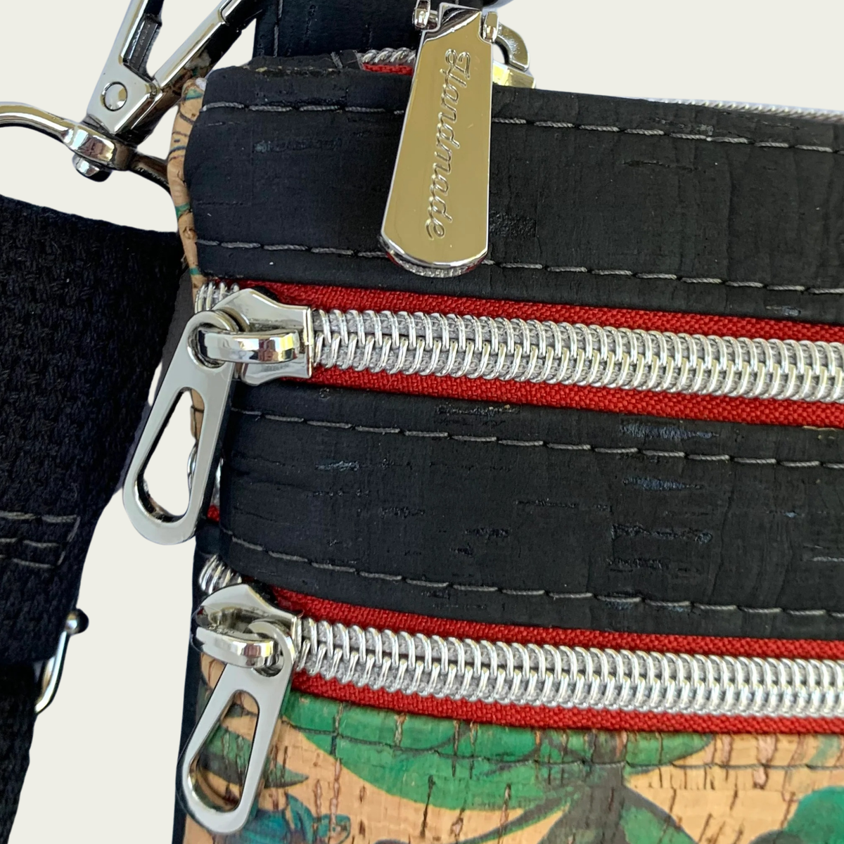 closeup of red zippers and nickel hardware on a black cork crossbody bag. “Handmade” is etched in the top zipper pull.