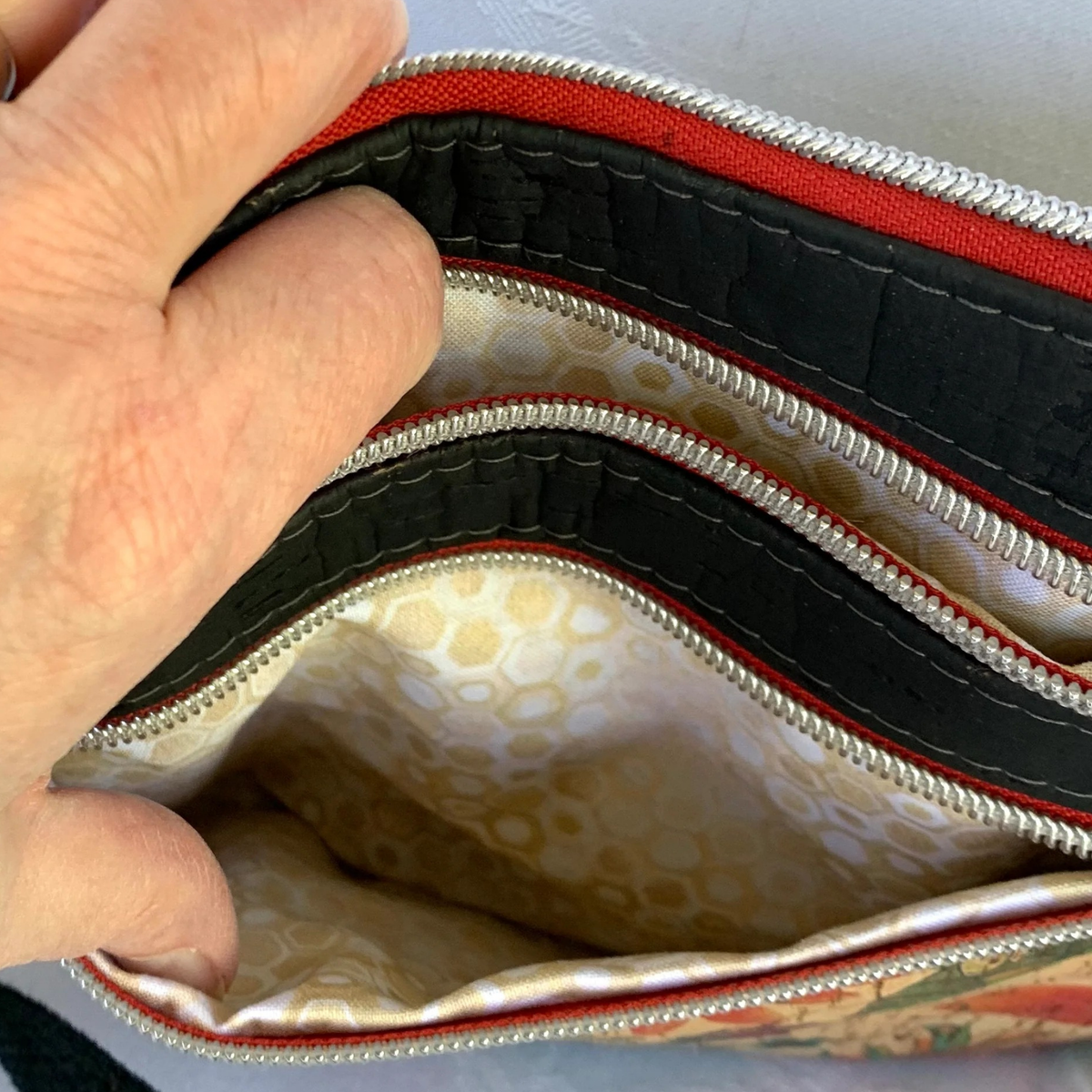 A closeup view of a cork handbag with 3 zippers. A hand is holding two of the zippers to pockets. The other zipper is closed on top of the bag. The pocket linings are a small geometric print in tan, gold, and white.