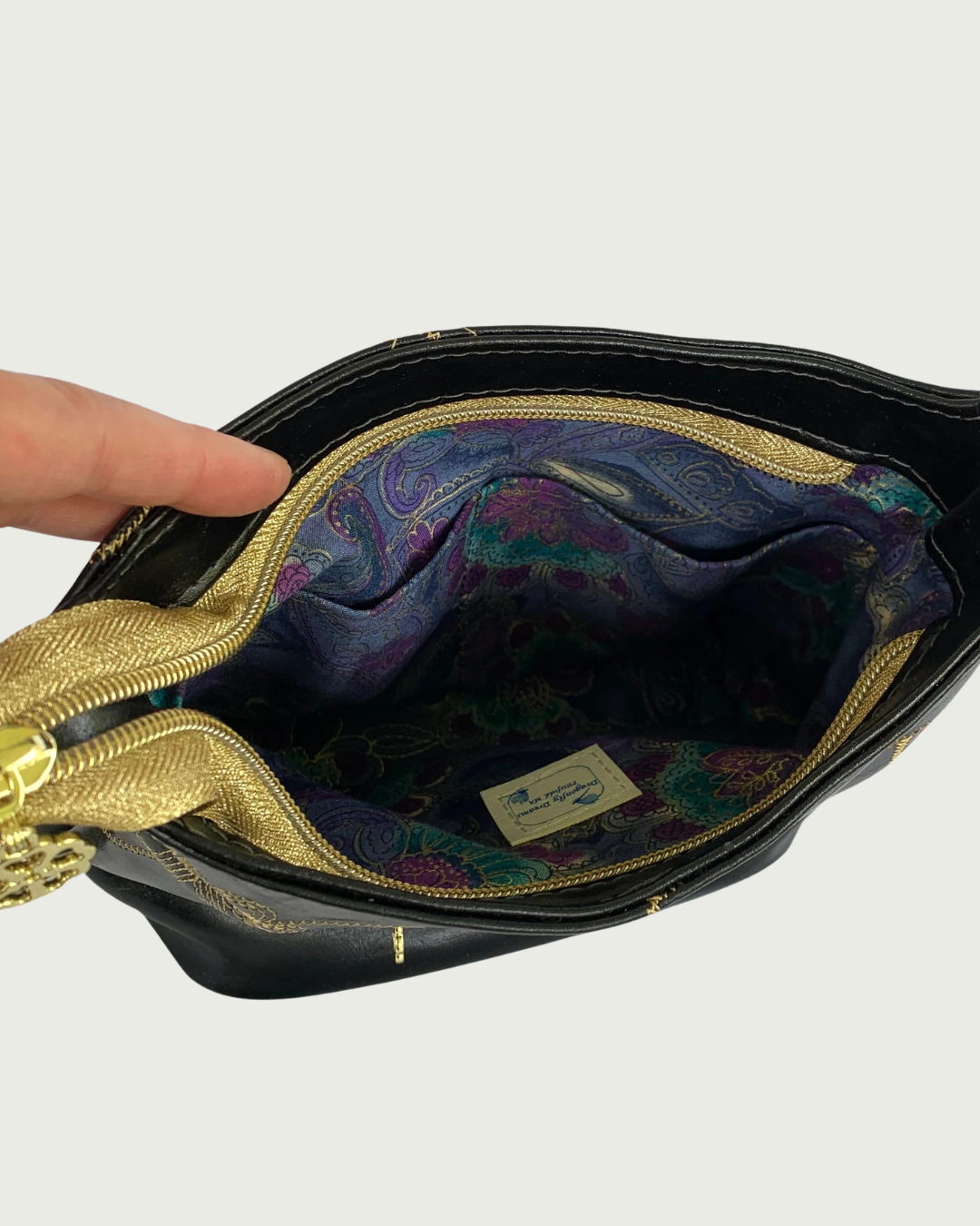 Handcrafted black vinyl slouchy shoulder bag with view of the interior showing an opened gold zippered closure. The lining is a rich, multicolor floral pattern with gold thread running through. There are two slip pockets and a label for Dragonfly Dreams sewn in.