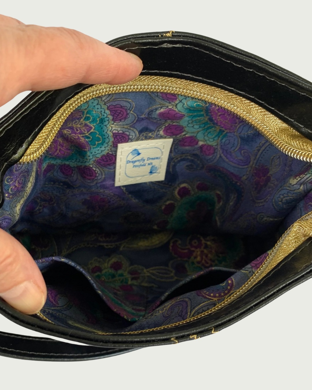 Handcrafted black vinyl slouchy shoulder bag with closeup view of a hand holding open a gold zipper showing the interior lining of a rich, multicolor floral pattern with gold thread running through. There are two slip pockets and a label for Dragonfly Dreams sewn in.