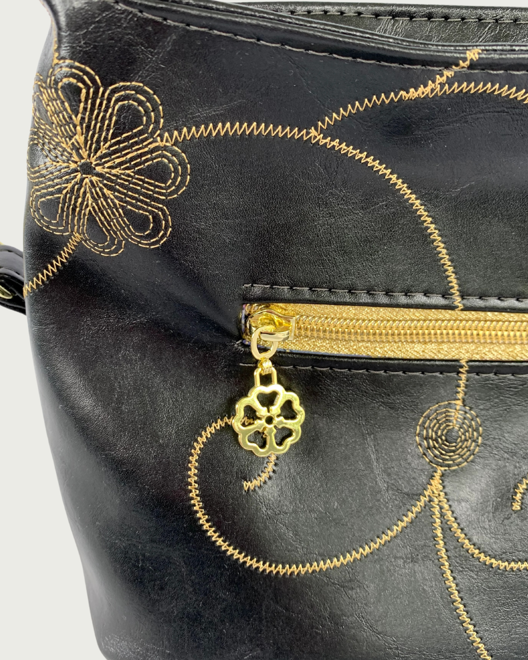 Handcrafted black vinyl slouchy shoulder bag with closeup of decorative gold stitching, gold hardware, detachable strap, and a small exterior pocket with gold zipper and zipper pull resembling the design of the bag.
