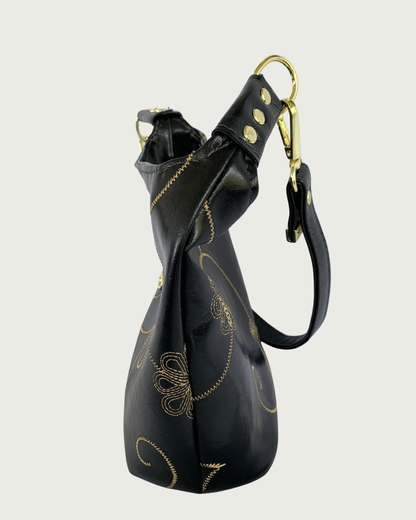 Handcrafted black vinyl slouchy shoulder bag side view with decorative gold stitching, detachable strap, and gold hardware including studs or rivets.