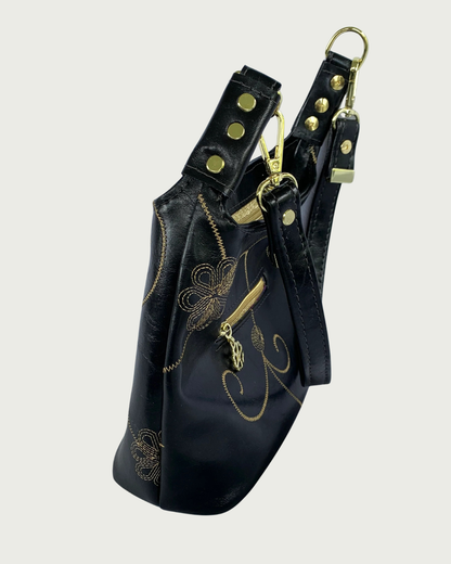 Handcrafted black vinyl slouchy shoulder bag side view with decorative gold stitching, exterior zippered pocket, detachable strap, and gold hardware.