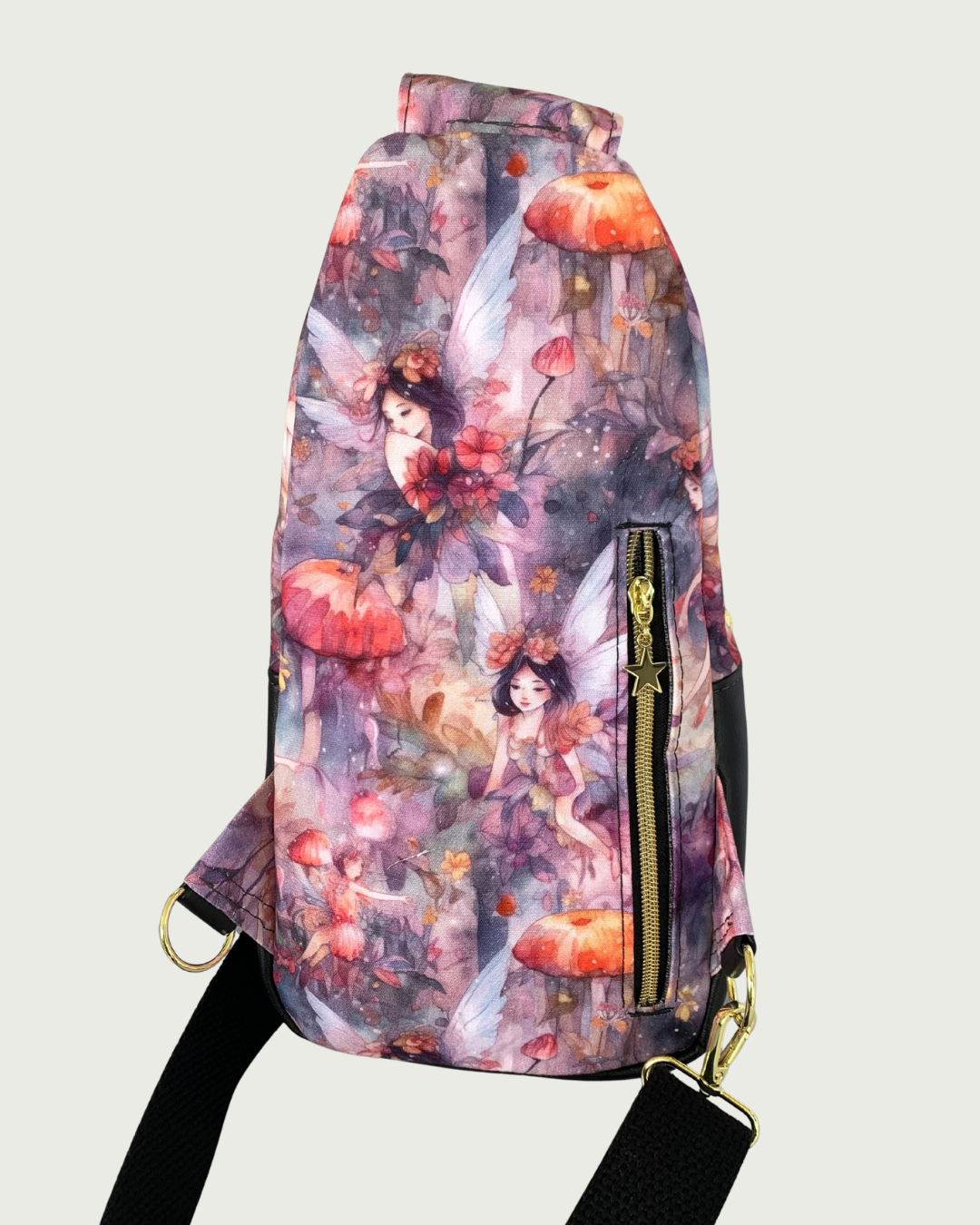Back view of handcrafted sling bag with a print of an enchanted forest with fairies and mushrooms in pinks and purples, grays and oranges. Water-resistant canvas bag with an adjustable wide black cotton webbing strap also repositional for left and right handed wear. All hardware is gold including the zipper teeth on black zipper tape that is positioned vertically for a concealed pocket.