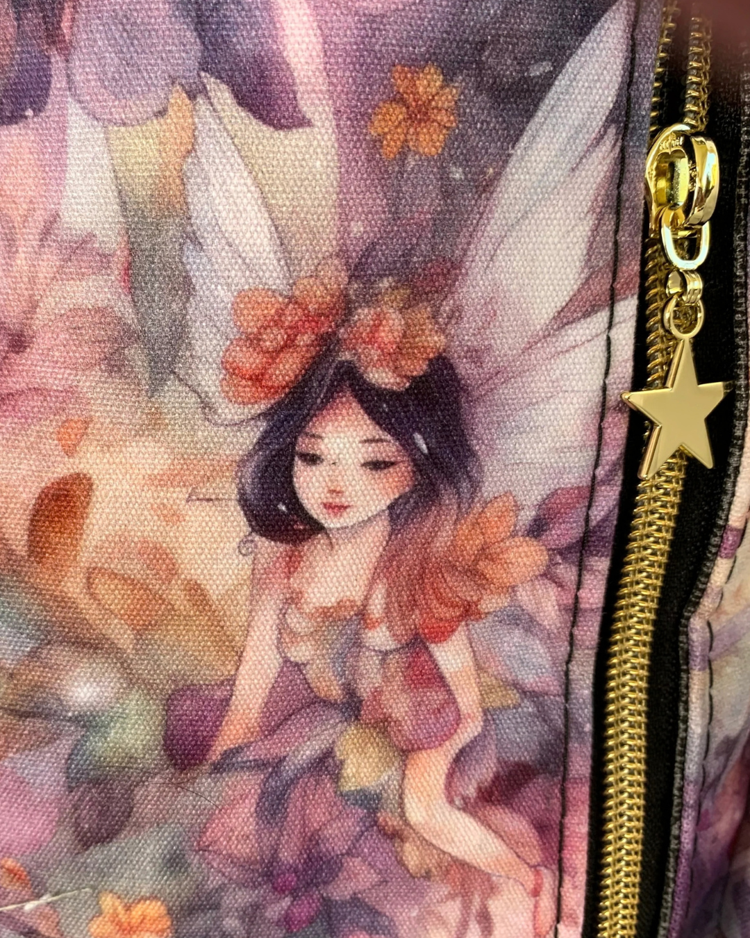 Back view closeup of handcrafted sling bag with a print of angels and flowers in pinks, oranges, purples and grays on water-resistant canvas. A vertical zipper is shown with black zipper tape, gold zipper teeth, and a gold star pull.