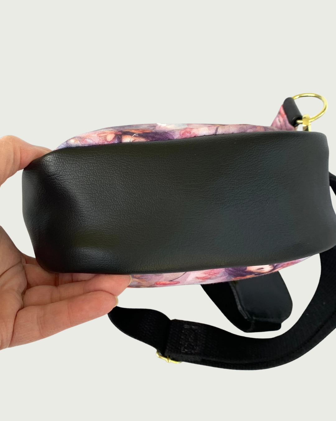 The bottom view of a handcrafted sling bag of water-resistant canvas in pinks, purples and grays. The bottom is also part of the gusset and it is made from black faux leather. An adjustable wide black cotton webbing strap is shown. All hardware is gold.