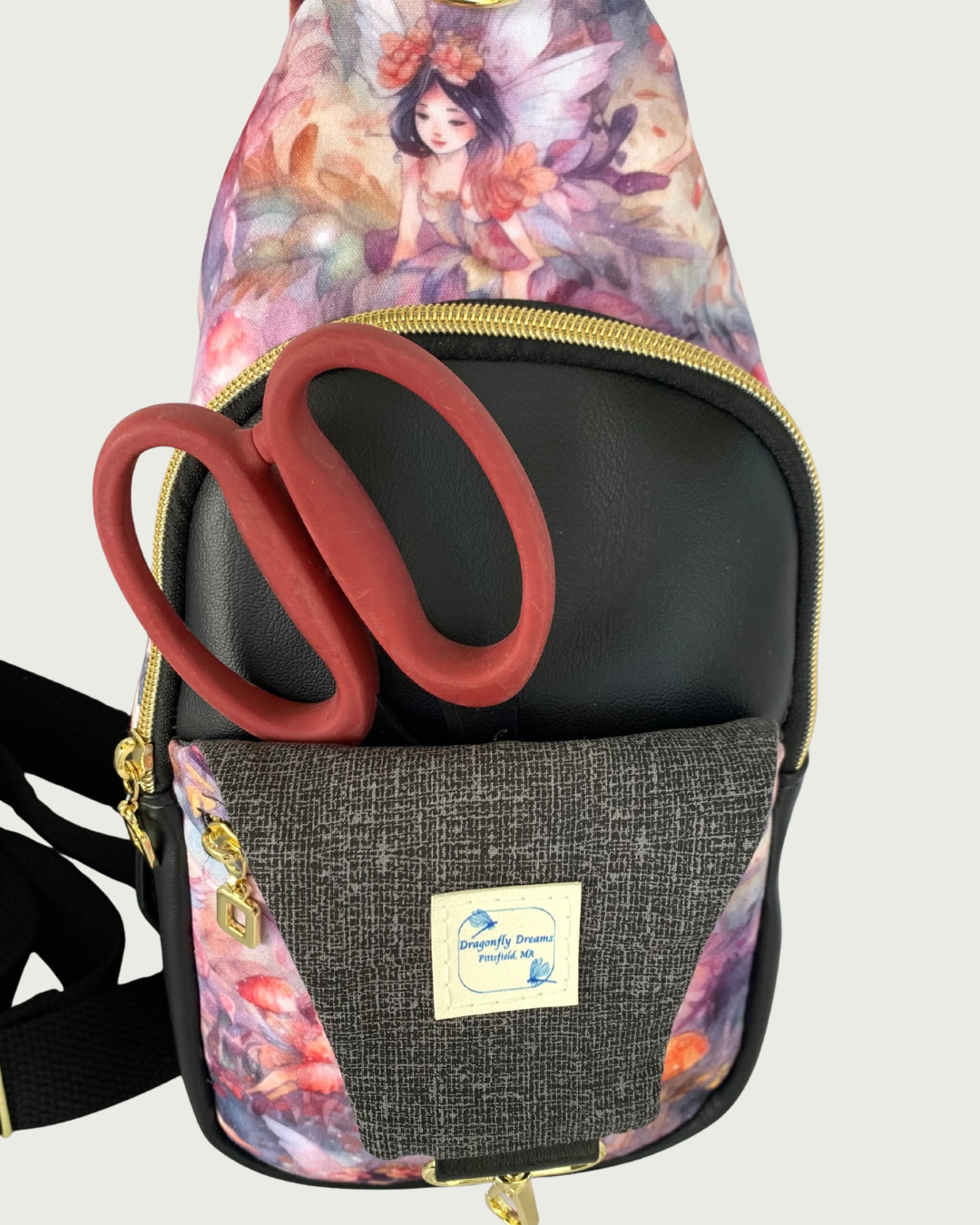 Handcrafted sling bag with a print of an enchanted forest with fairies and mushrooms in pinks and purples, grays and oranges. Water-resistant canvas bag with black faux leather accents and a black wide, adjustable cotton webbing strap. A pair of red-handled scissors is in the pocket space between an open panel of charcoal and light gray crosshatch lining and the main compartment of black faux leather vinyl. The compartment is zipped closed with black zipper tape and gold teeth. A Dragonfly Dreams label is s