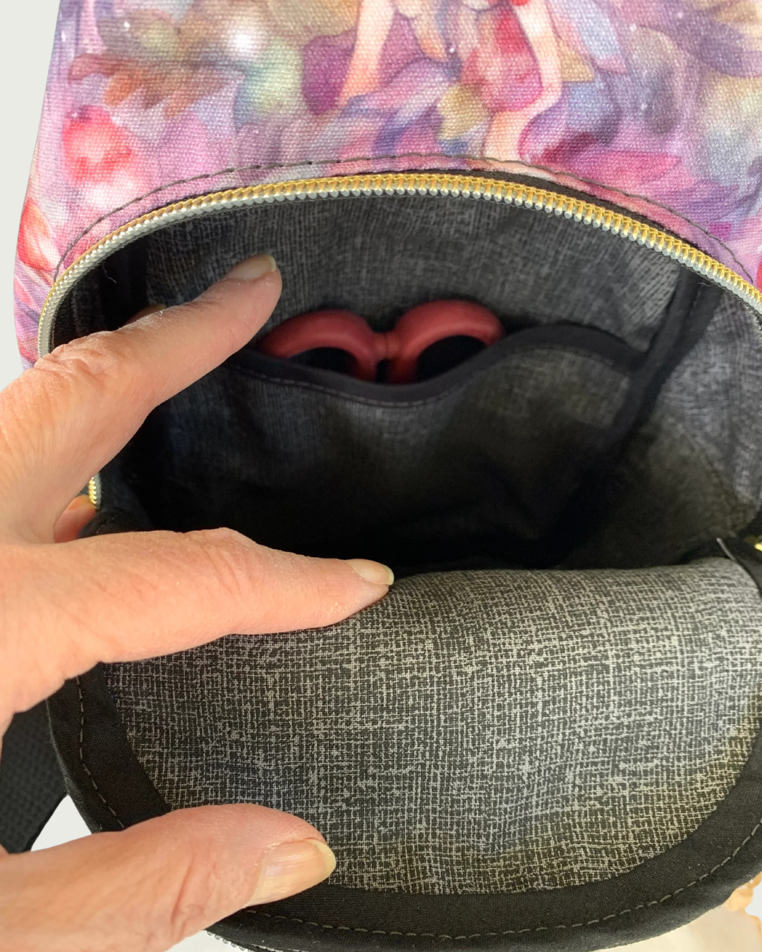 Handcrafted sling bag with a print of pinks, purples, grays on water-resistant canvas. A hand is holding open the main compartment revealing a lining of charcoal and light gray crosshatch with black binding on the seams. The interior slip pocket has a pair of red-handled scissors peeking through.