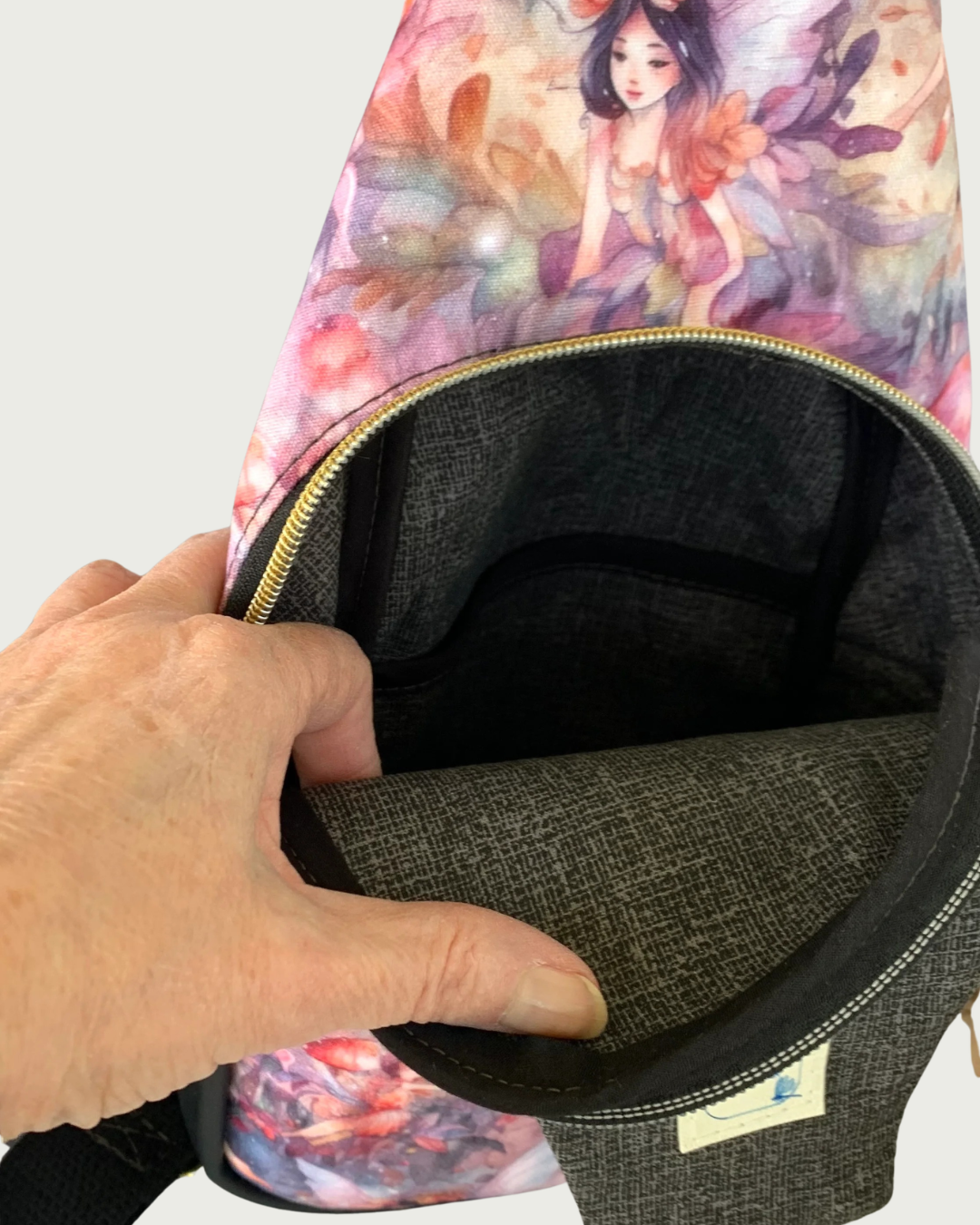 Handcrafted sling bag with a print of an enchanted forest of fairies on water-resistant canvas. A hand is holding open the main compartment revealing a lining of charcoal and light gray crosshatch, a slip pocket, and black binding on the seams.