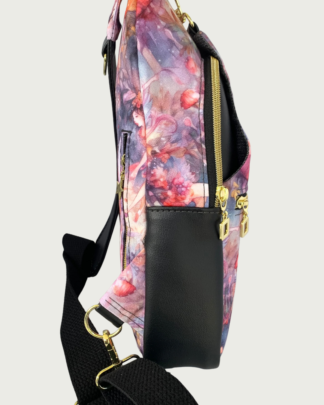 Handcrafted sling bag standing sideways of water-resistant canvas with a print of an enchanted forest with fairies and mushrooms in pinks and purples, grays and oranges. Black faux leather accents with an adjustable wide black cotton webbing strap. All hardware is gold including the zipper teeth on black zipper tape on the front pocket as well as the main compartment. A front panel that has the pocket unhooks with a swivel held onto a d-ring. Behind that is a curved zippered main compartment made of the bla