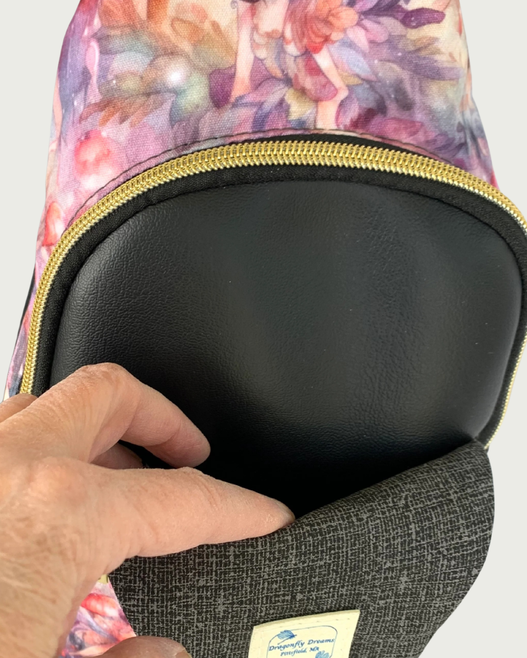 Handcrafted sling bag with a print of an enchanted forest with fairies and mushrooms in pinks and purples, grays and oranges. Water-resistant canvas bag with black faux leather accents. A hand is holding open a slip pocket in the space between an open panel of charcoal and light gray crosshatch lining and the main compartment of black faux leather vinyl. The compartment is zipped closed with black zipper tape and gold teeth. A Dragonfly Dreams label is sewn into the lining.